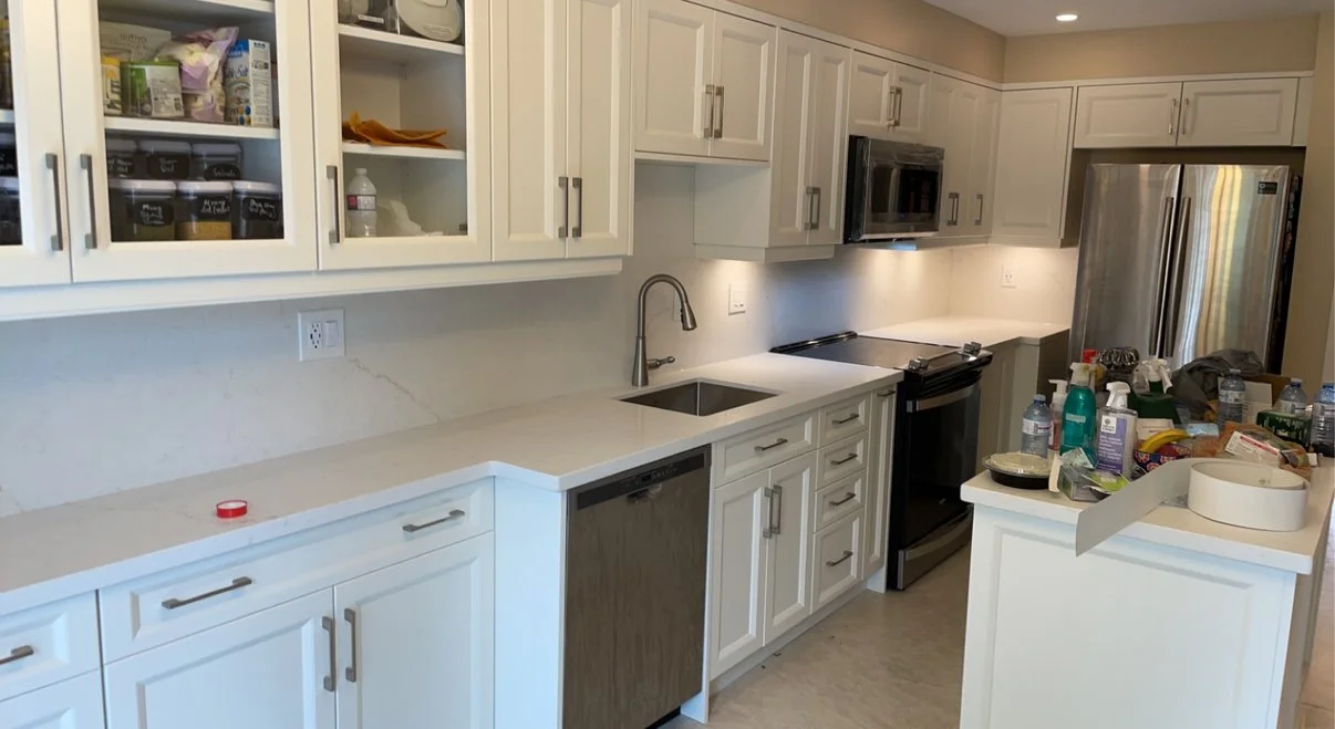A modern kitchen with white cabinets, a gray marble backsplash, and stainless steel appliances, including a refrigerator, microwave, and oven. The countertop has clutter such as water bottles, food containers, and miscellaneous items.