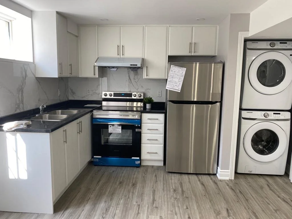 A modern kitchen with white cabinets, black countertops, stainless steel appliances including a refrigerator, oven, and microwave, and a stacked washer and dryer.