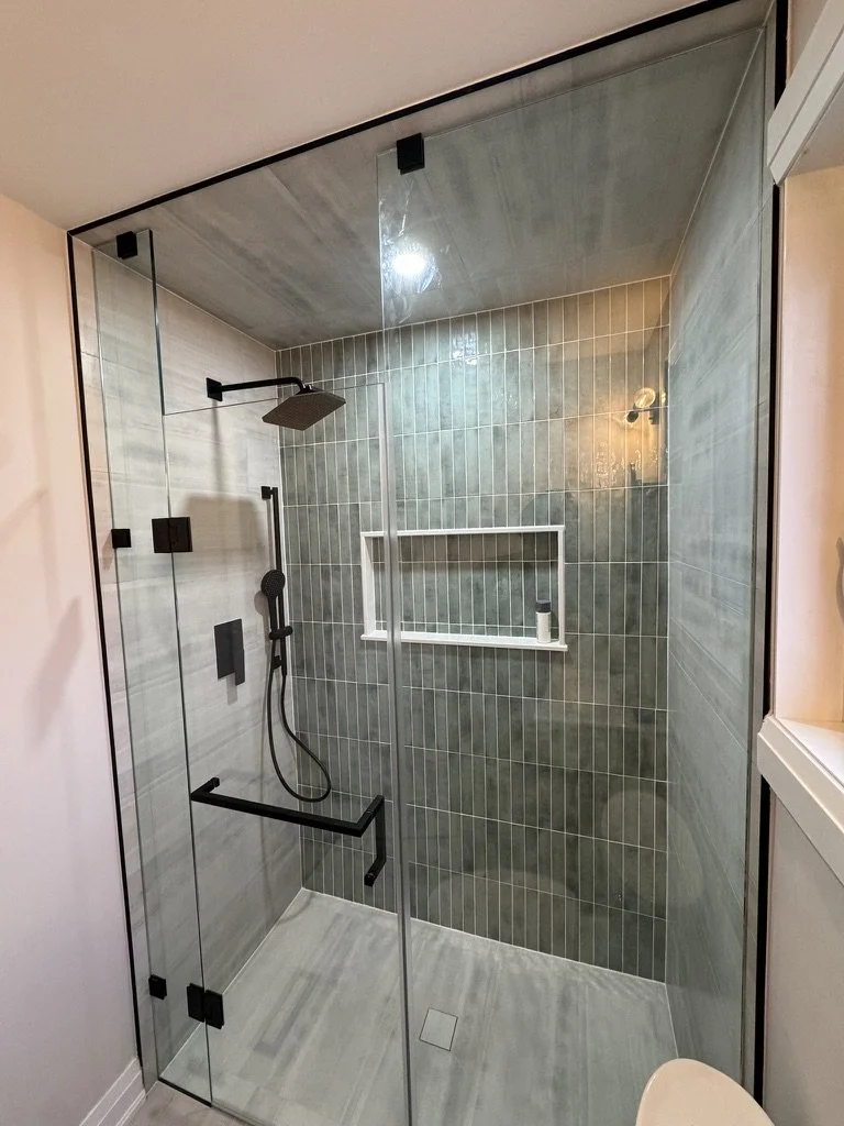 Modern walk-in shower with glass enclosure, black fixtures, rainfall showerhead, handheld shower, built-in shelf, grey tile walls, and a light-colored tile floor.