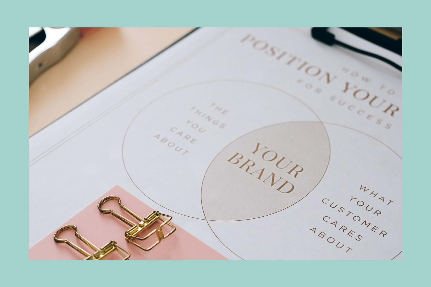 A printed worksheet titled 'How to Position Your Brand for Success' with overlapping circles labeled 'The Things You Care About,' 'What Your Customer Cares About,' and 'Your Brand.' A pink notepad with gold binder clips is at the bottom of the worksheet, and a black pen is in the top right corner.