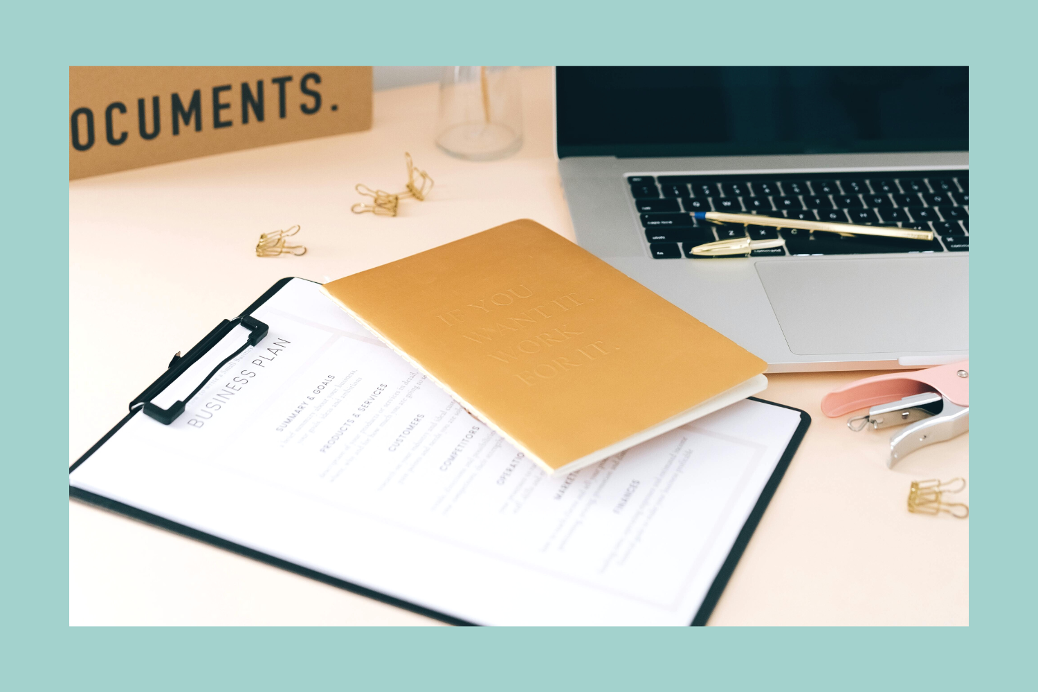 A workspace desk with a laptop, a clipboard with a business plan, a gold notebook, paper clips, and a pink and gold stapler.