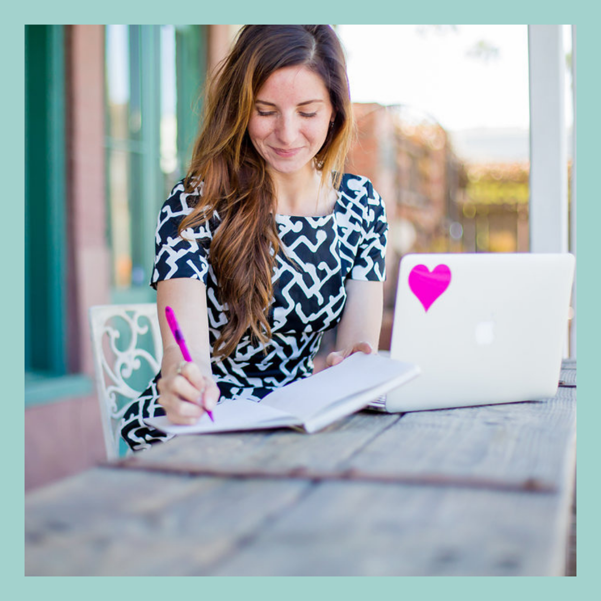 A woman with long brown hair writing in a notebook, sitting at a table with a silver laptop that has a pink heart sticker on it, near a large window with a blurred outdoor background.