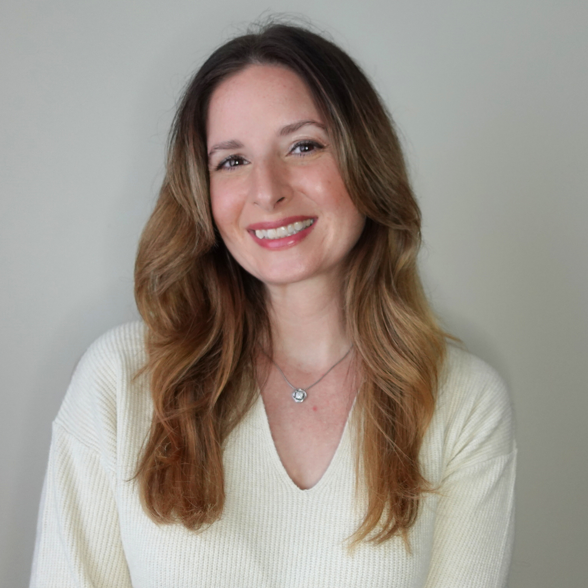 Portrait of a woman with long wavy brown hair wearing a light-colored top and a necklace, smiling at the camera against a plain background.