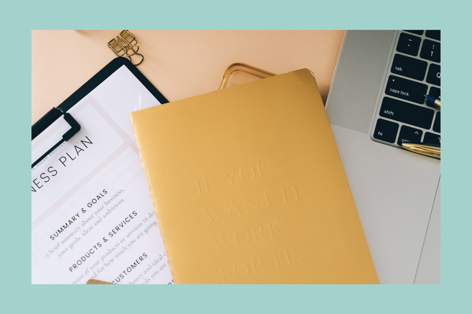 Desk workspace with a laptop, a gold notebook, a business plan document, and a gold paper clip on a teal background.