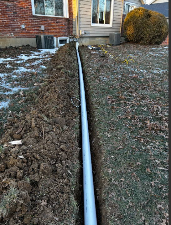 A large trench dug along the side of a house with a pipe installed in it, surrounded by dirt and grass with some patches of snow.