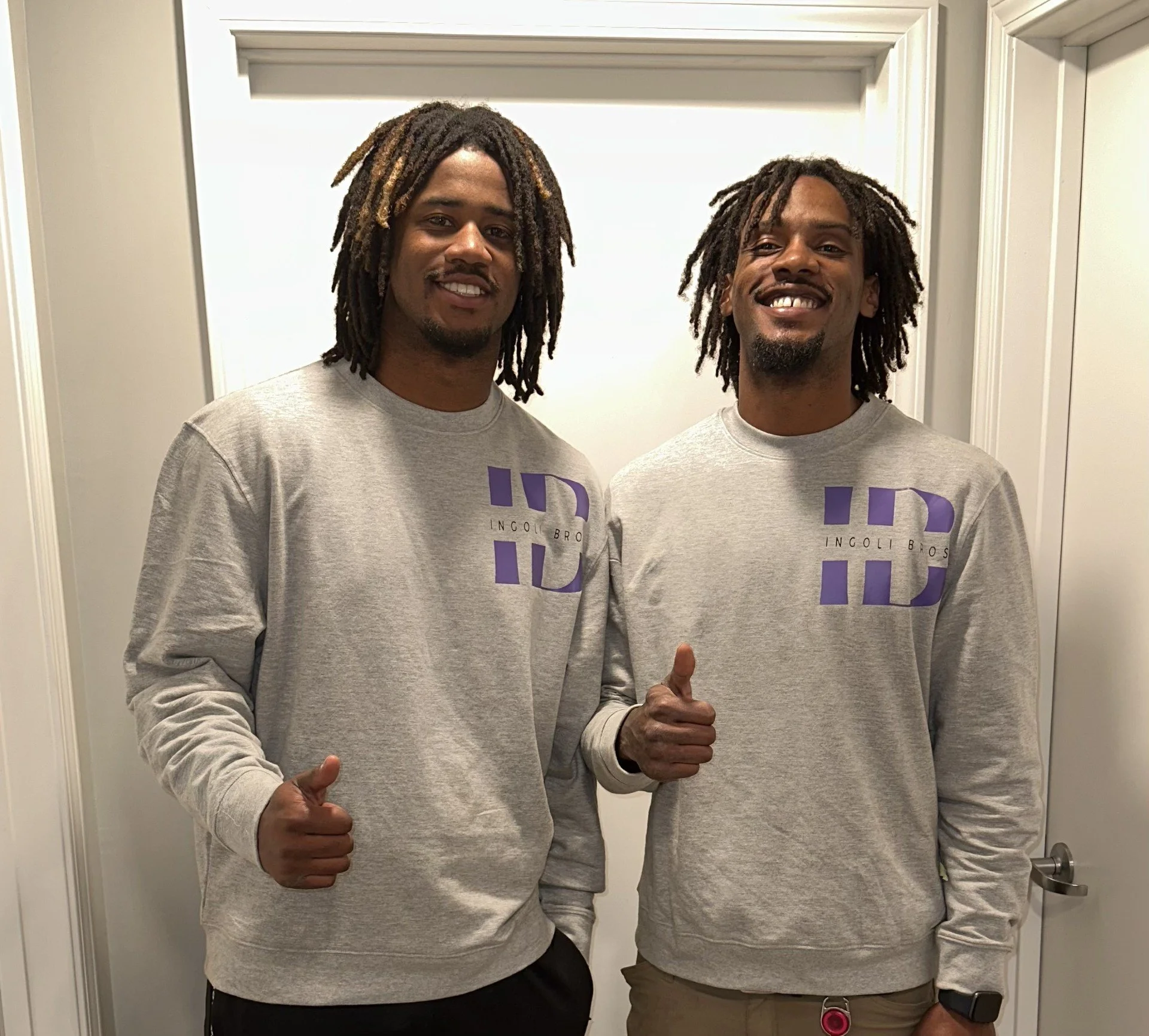 Two men with dreadlocks wearing matching gray sweatshirts with purple 'ID' logos, standing indoors in front of a white door, smiling, giving thumbs-up gestures.