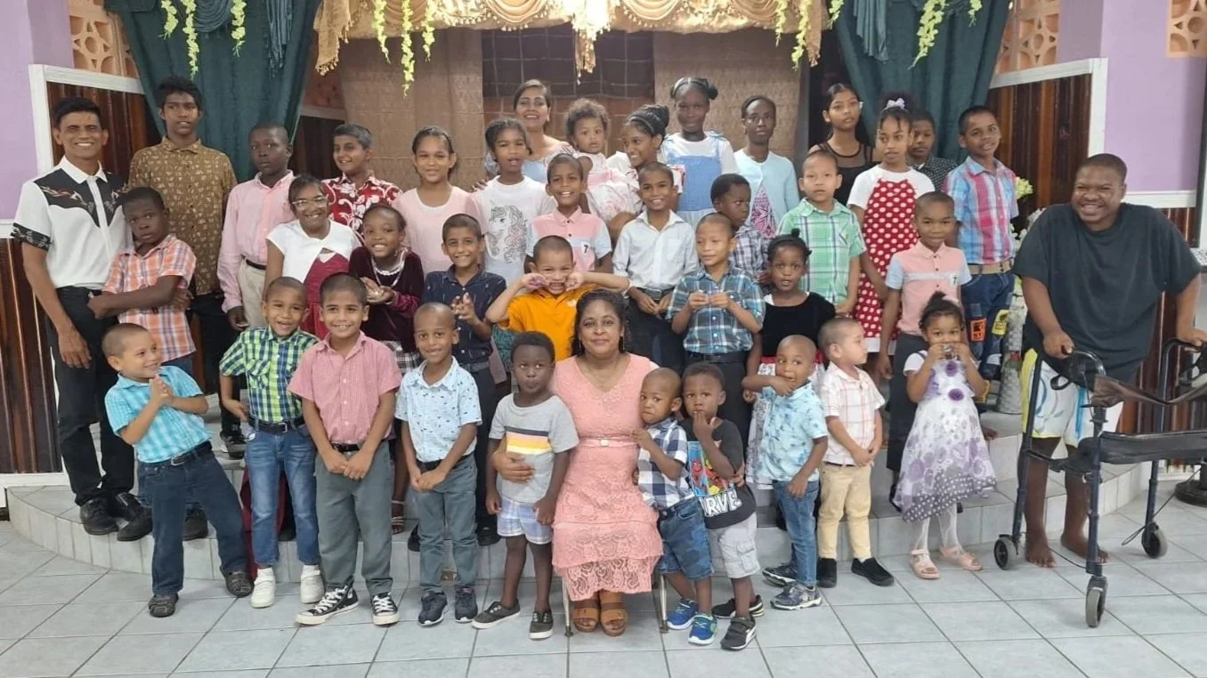 A large group of children and two adults posing indoors, with children smiling and some using a walker for assistance.