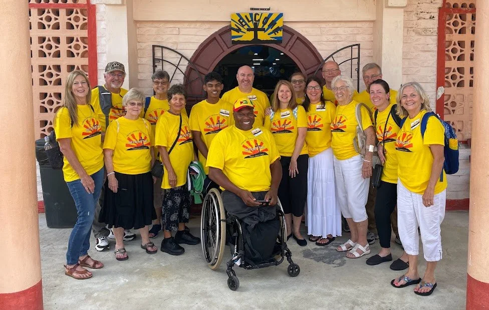 Group of people wearing matching yellow shirts with a logo, standing outside a building entrance. One person in a wheelchair is in the front center.