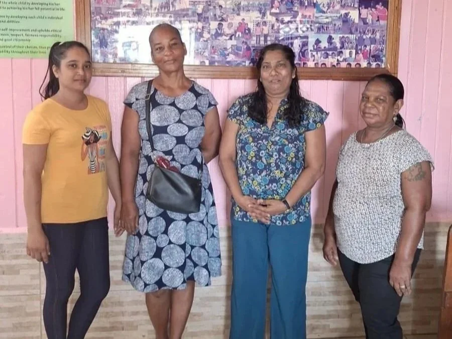 Four women standing indoors against a pink wall, with a collage of photos behind them.