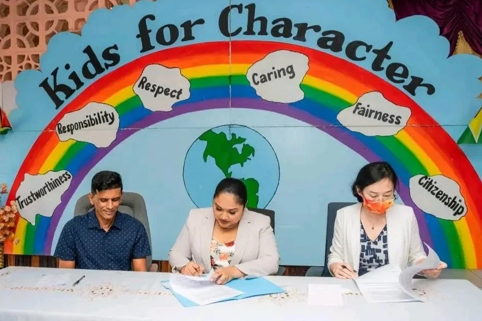 Three people seated at a table in front of a colorful rainbow-themed background that reads "Kids for Character." The background displays words like responsibility, respect, trustworthiness, caring, fairness, citizenship, and responsibility, with a globe illustration in the center.
