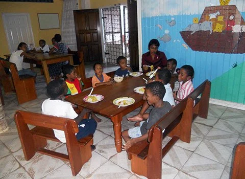 Group of children and an adult sitting around a table with plates of food in a room with colorful mural on the wall.