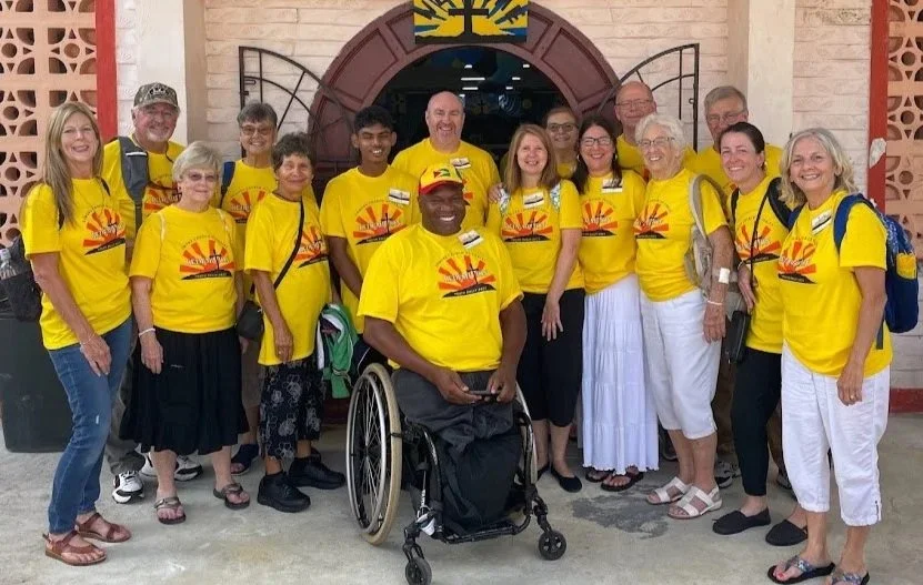Group of people wearing matching yellow t-shirts with sunrise logo, gathered outside a building with an arched entrance and a coat of arms above the door, smiling for a photo.