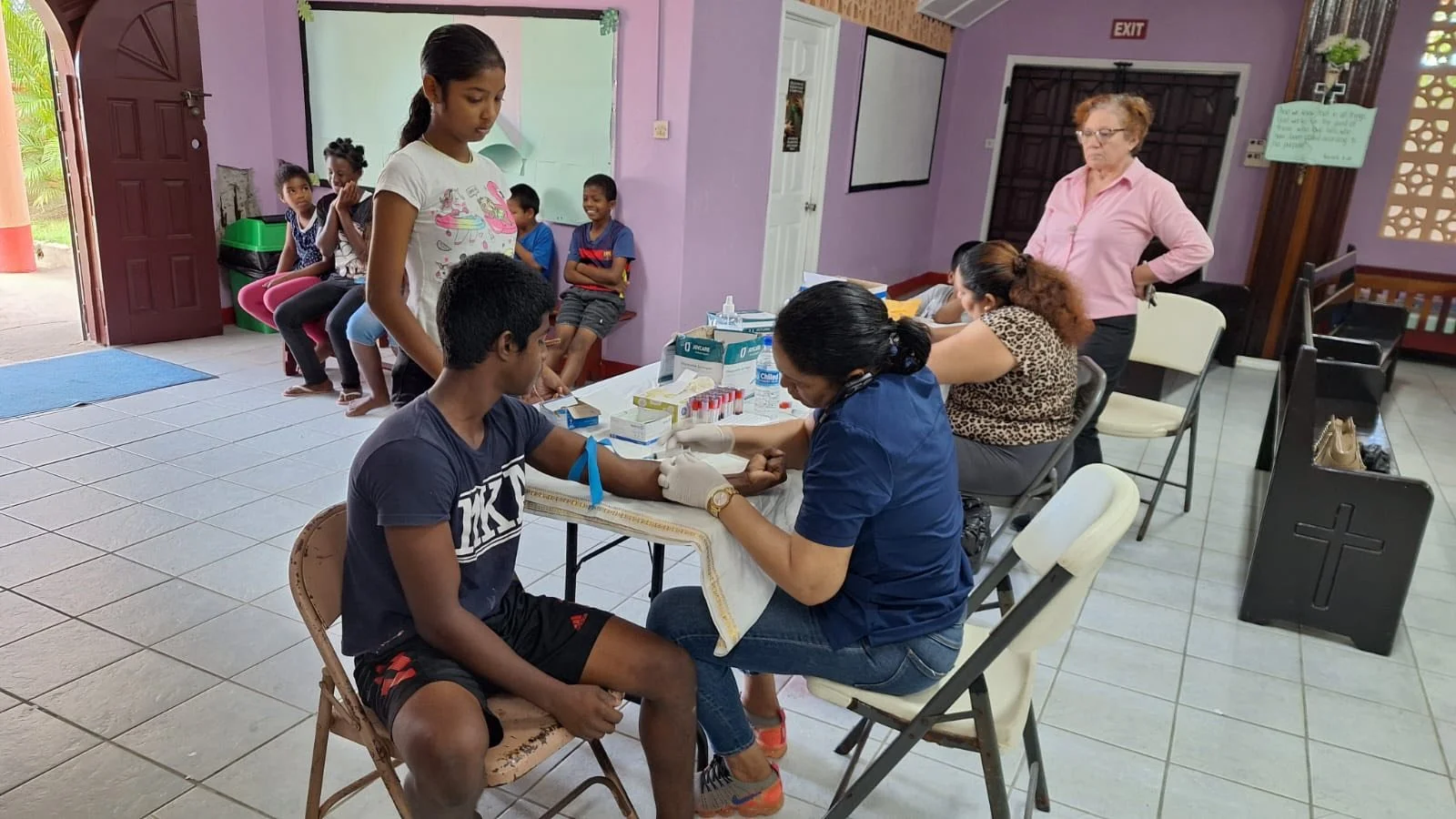 A group of children and adults at a medical vaccine clinic inside a room with purple walls. One child is sitting while a nurse administers a shot, and several children are waiting in line or sitting in the background.
