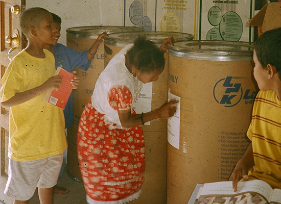 Children in a community center around large recycling bins, with one girl in a red dress writing on a bin and other children observing, playing, and holding books.