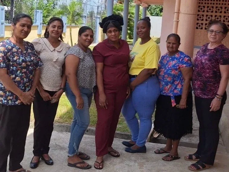 Group of seven women standing outdoors in casual clothing with a building and greenery in the background.
