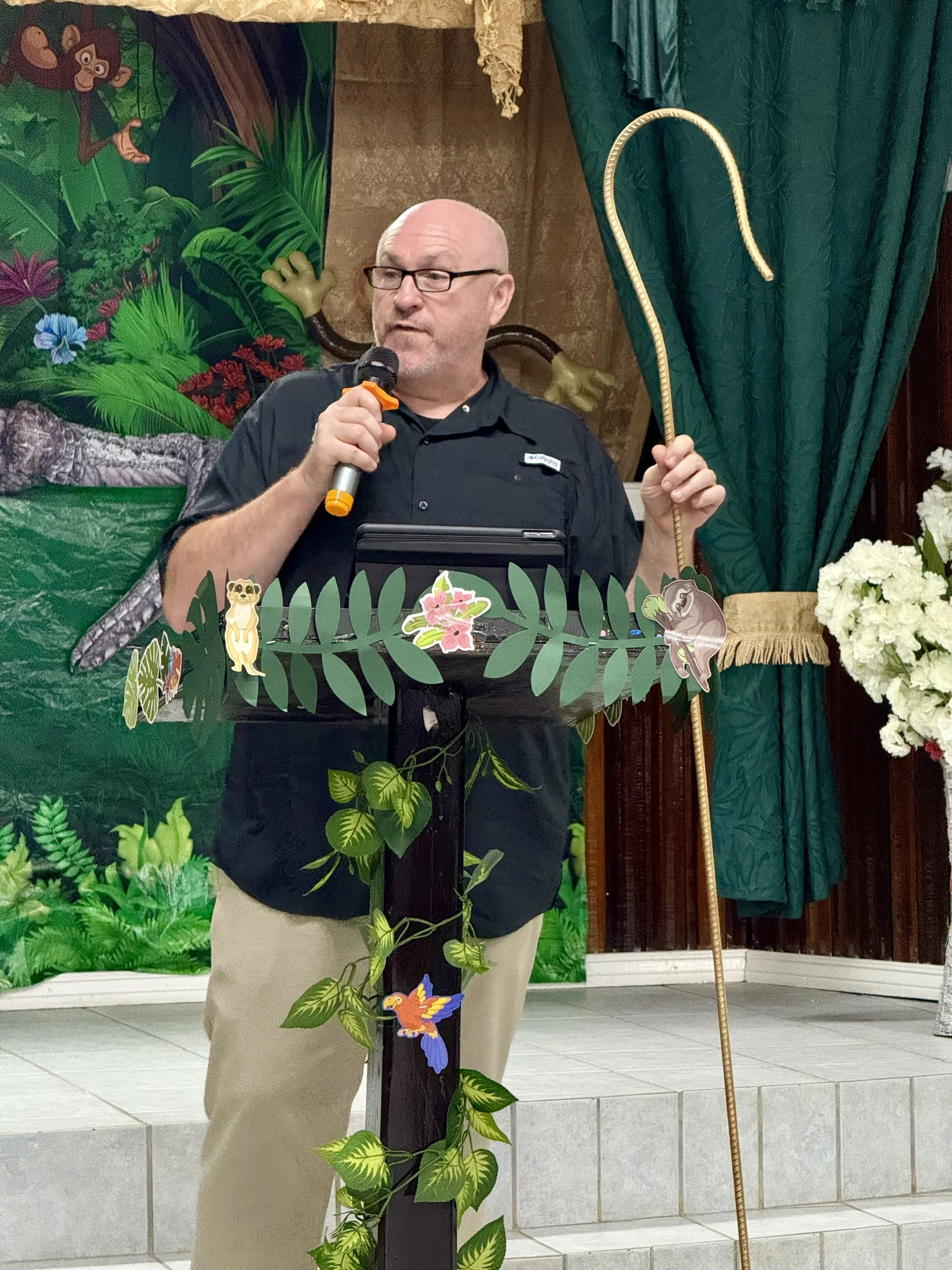 A bald man with glasses speaking into a microphone, holding a shepherd's crook, standing behind a decorated podium with jungle-themed cutouts, in front of a jungle-themed backdrop with green curtains and flowers.