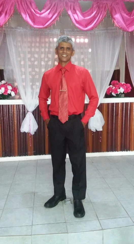 A man in a red shirt, black pants, and a red tie standing indoors in front of a decorated background with pink drapes and pink flowers.