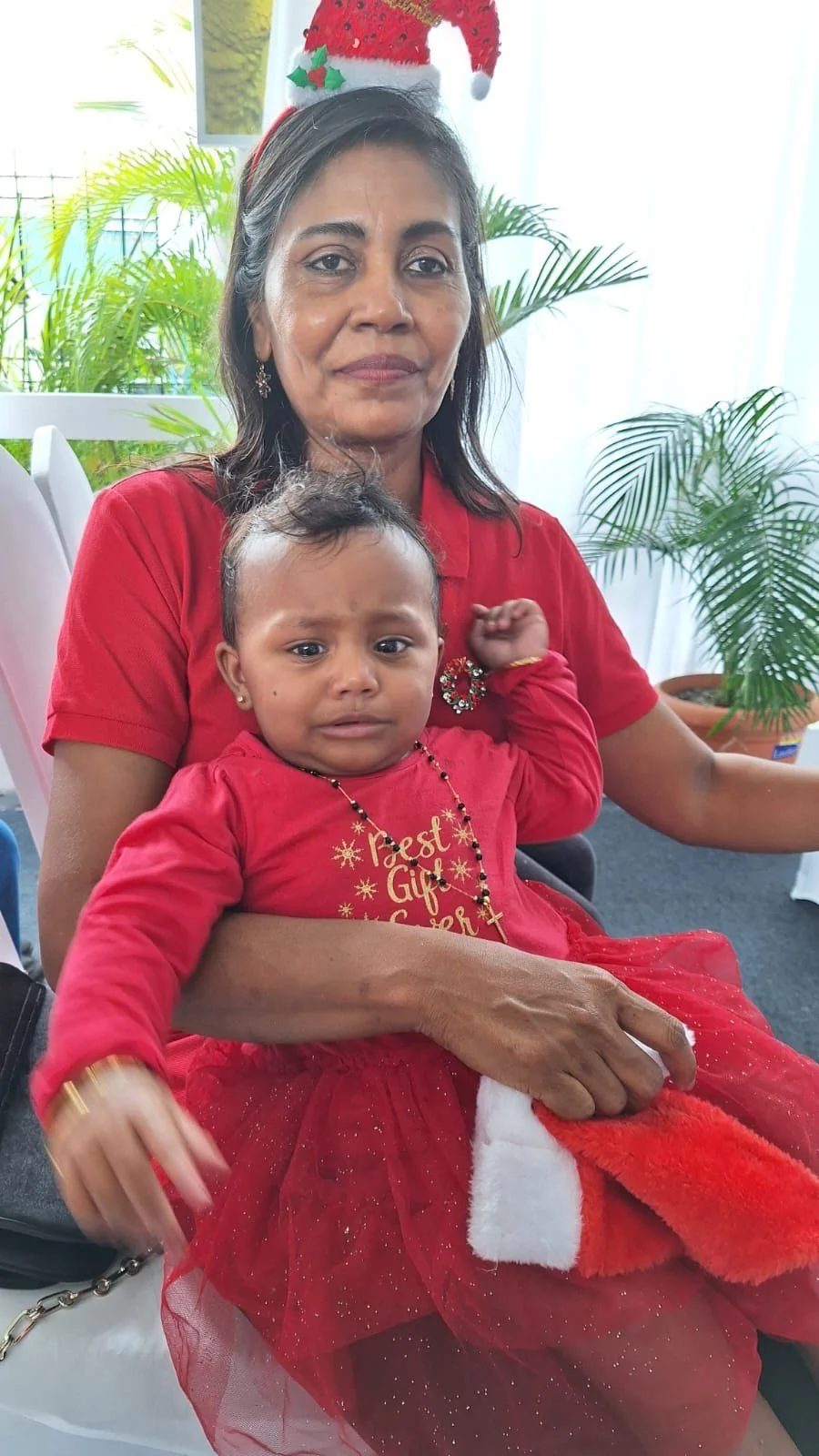 A woman and a young girl dressed in red, sitting indoors surrounded by green plants, with a festive holiday hat on the woman's head.