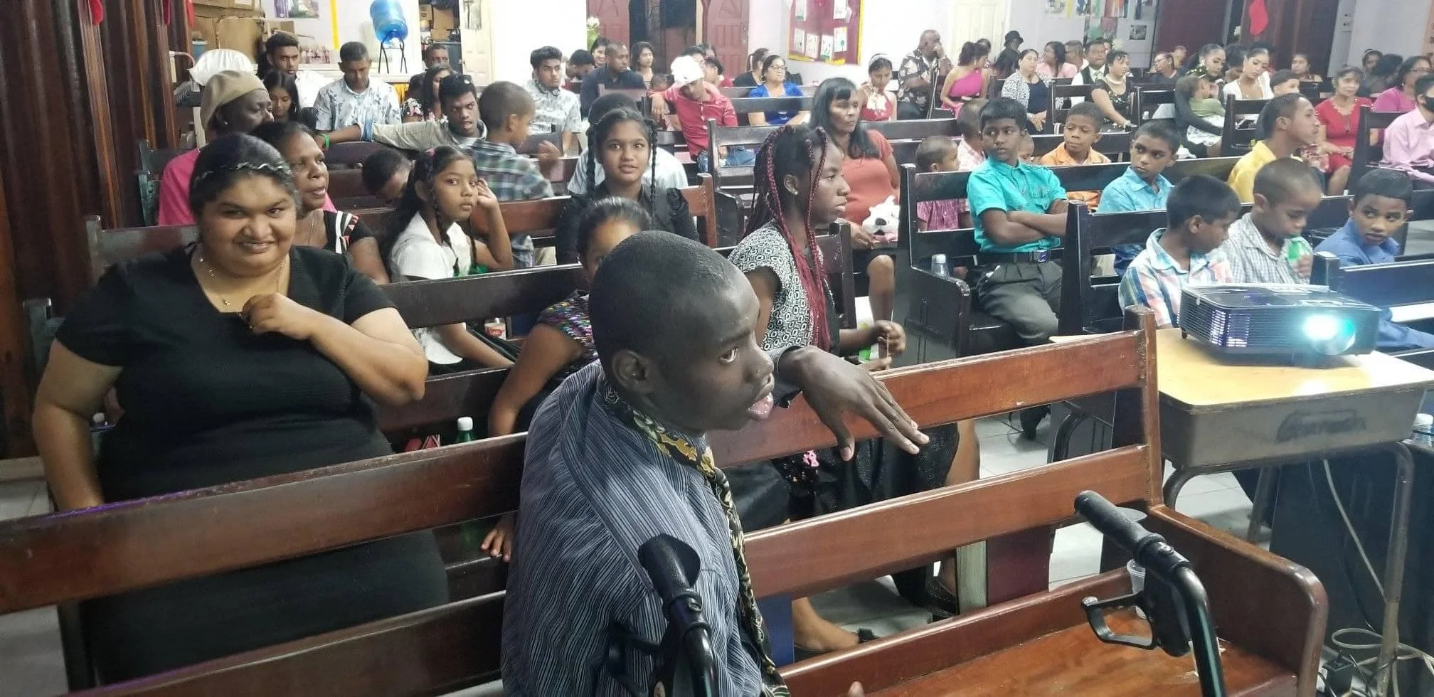 A group of diverse people, including children and adults, seated in a room attending a presentation or event, with a projector on a table at the front.