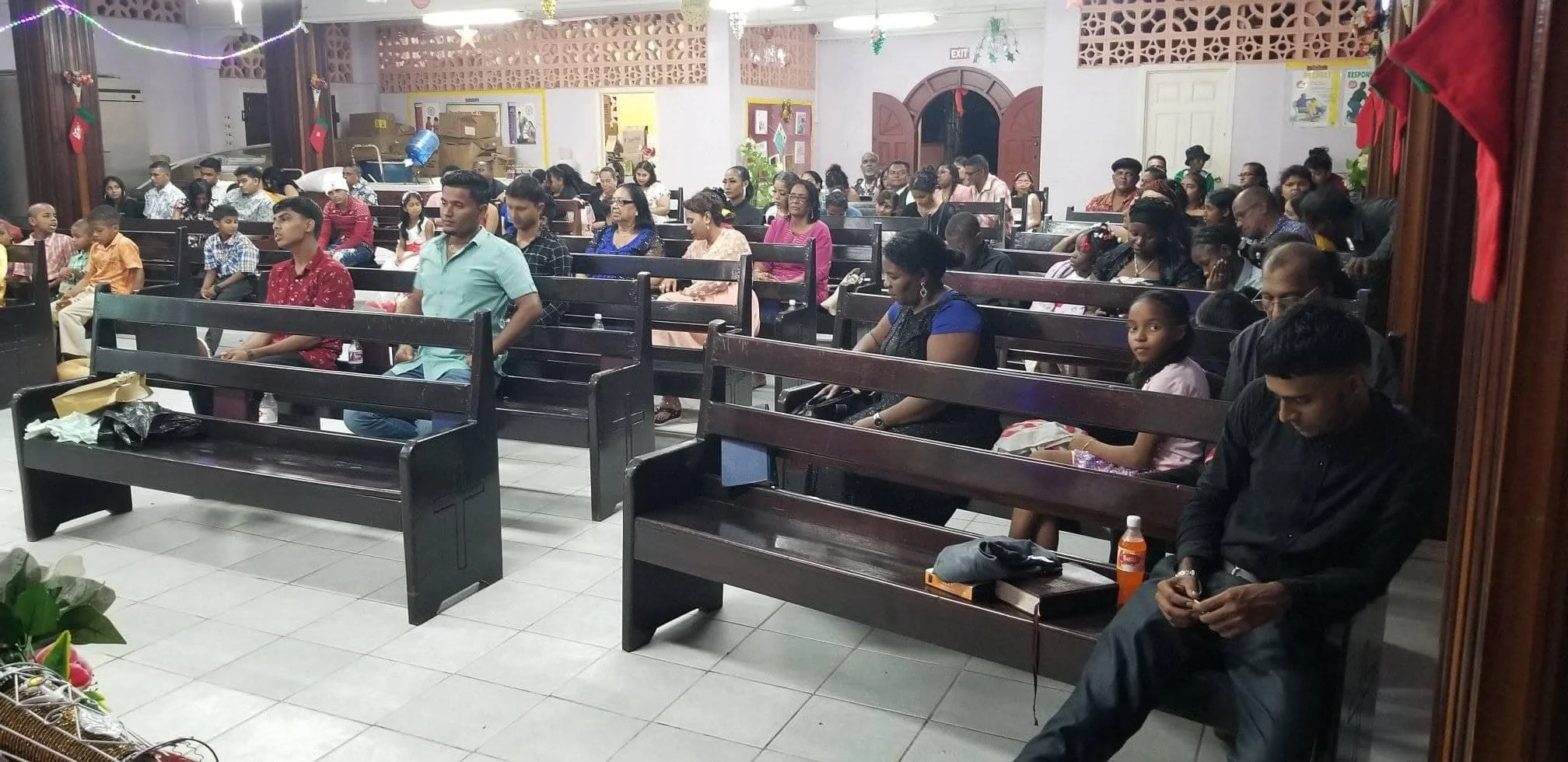 A group of adults and children seated in a church or hall, some are sitting with eyes closed, others are looking forward or using mobile devices. The room has dark wooden pews, holiday decorations, and a few gifts or packages on the seats.