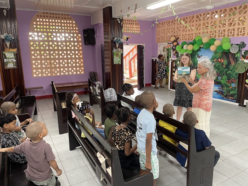 Children seated in pews in a decorated room with colorful balloons, a nature-themed backdrop, and two women at the front, one reading a book to the group.