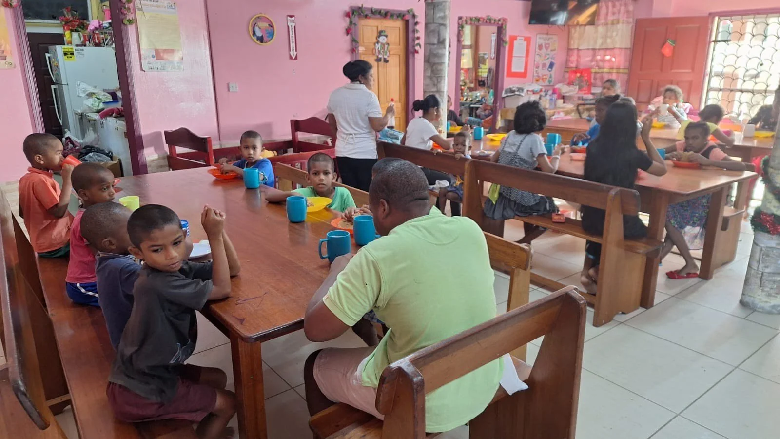 Children and adults sitting at wooden tables in a colorful room, eating and engaging in activities, with a school or daycare setting.