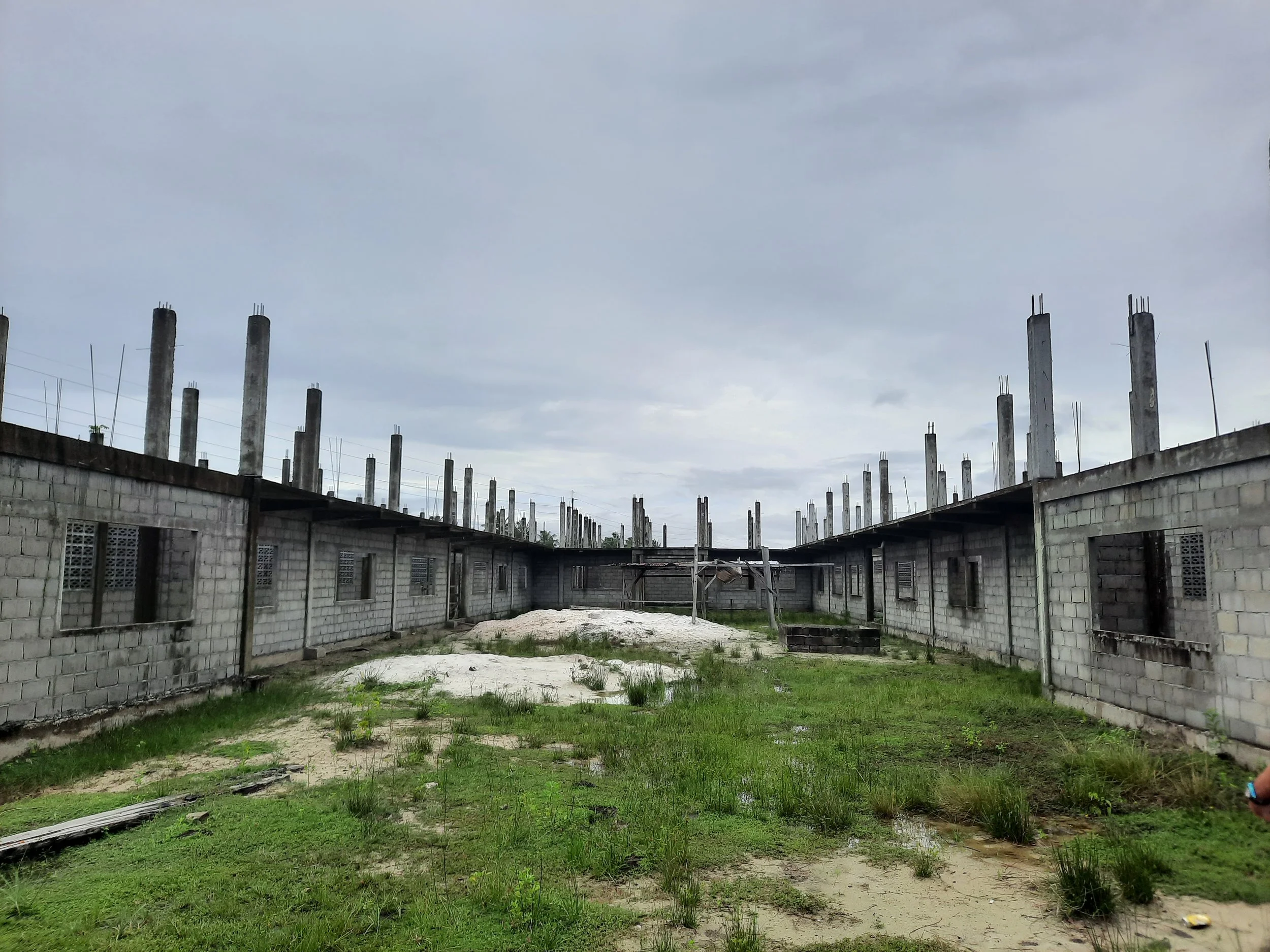 An under-construction building with gray concrete block walls and tall vertical concrete pillars, some with exposed steel rebar, on a grassy area under a cloudy sky.