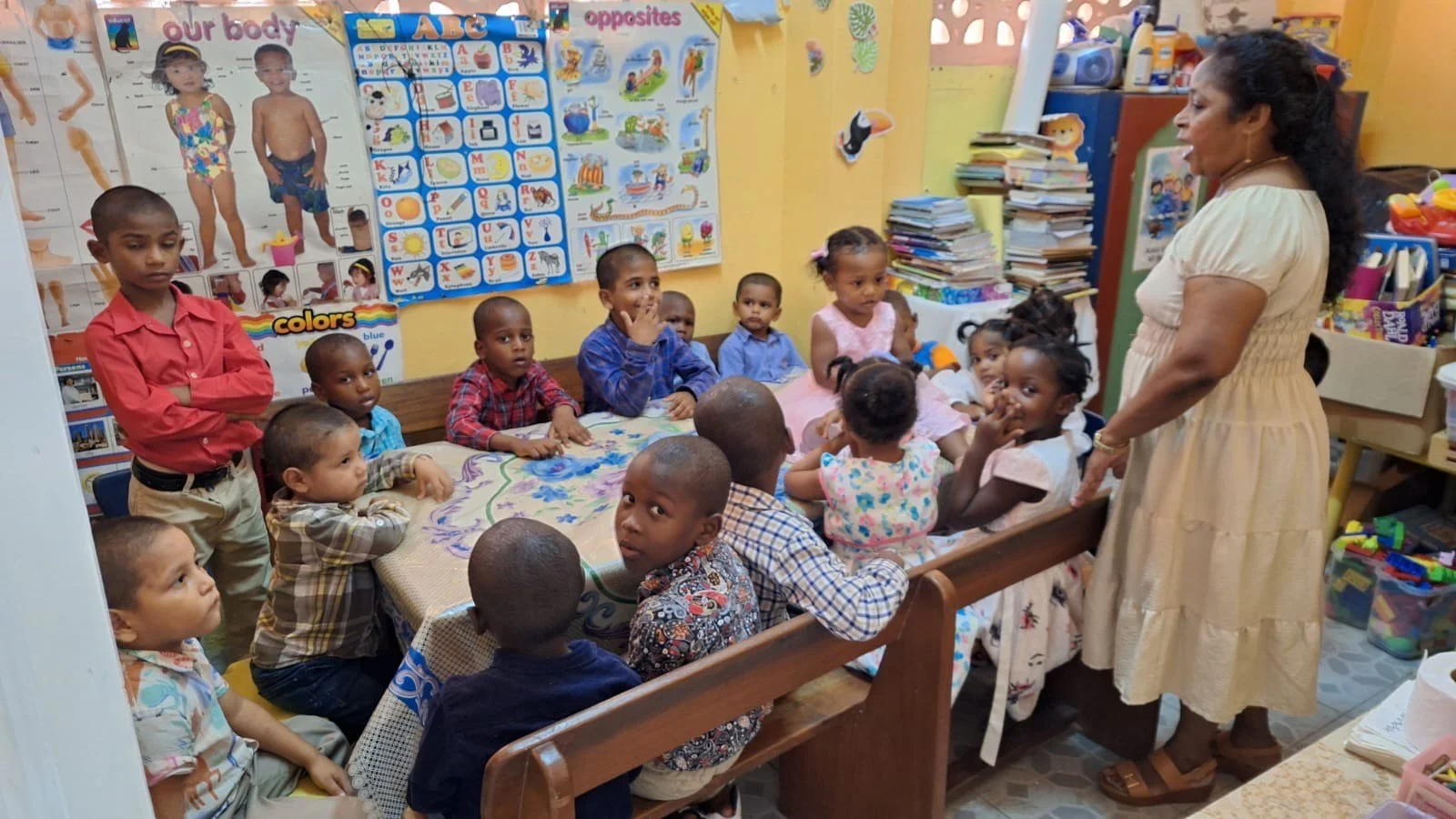 A teacher standing in front of a classroom filled with young children sitting on a bench and at a table, engaging during a lesson. The classroom has colorful educational posters on the wall, including ones about body parts and opposites.