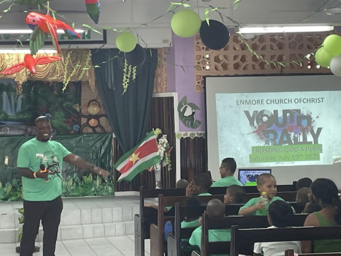 A man in a green shirt holding a flag of South Sudan, speaking to a group of children in a church setting with themed decorations and a projector screen displaying 'Youth Day'