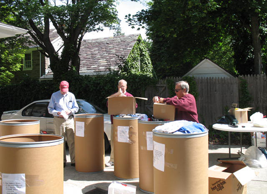 Three people are outdoors at a garage sale, sorting through large cardboard storage bins with various items. There are tables with more items and a blue car parked in the background, with trees and a house nearby.