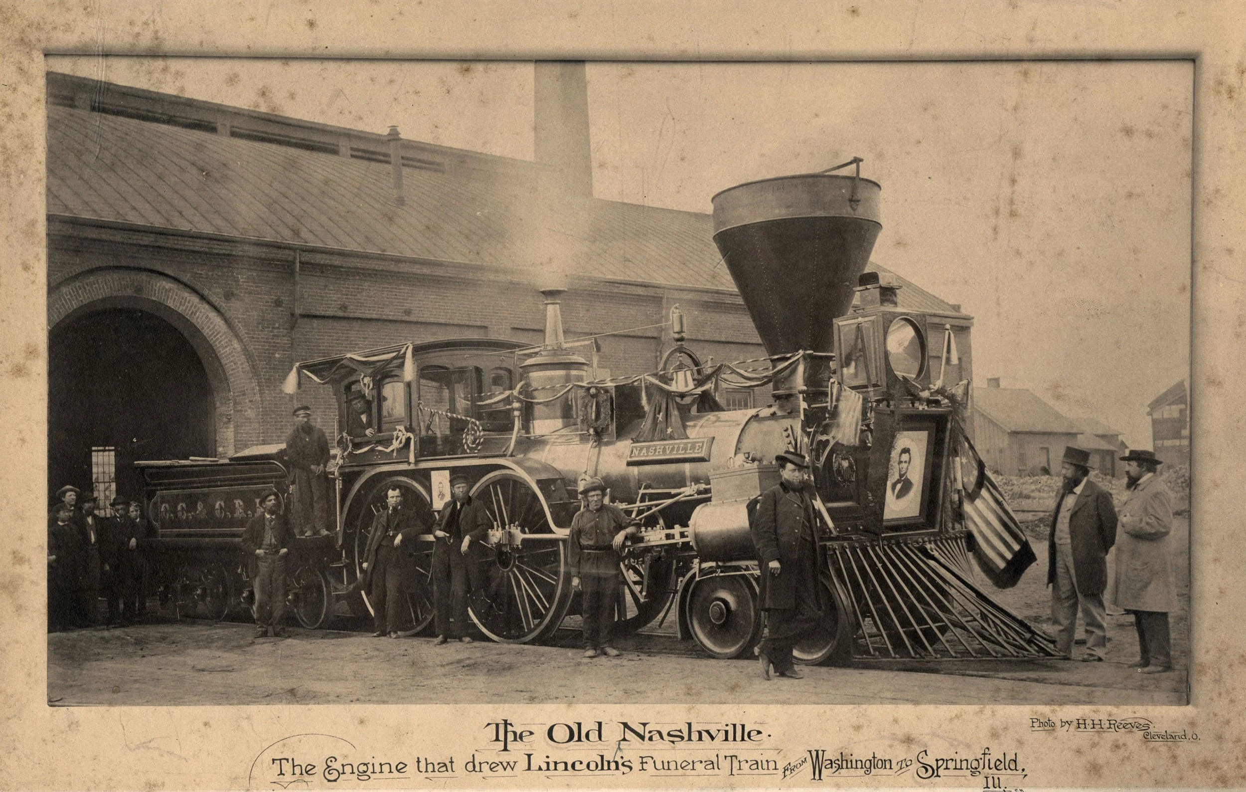 Black and white photograph of a vintage steam locomotive with a large funnel, labeled 'Nashville,' surrounded by men in period clothing, in front of an old brick building with a curved arch entrance, during daytime.