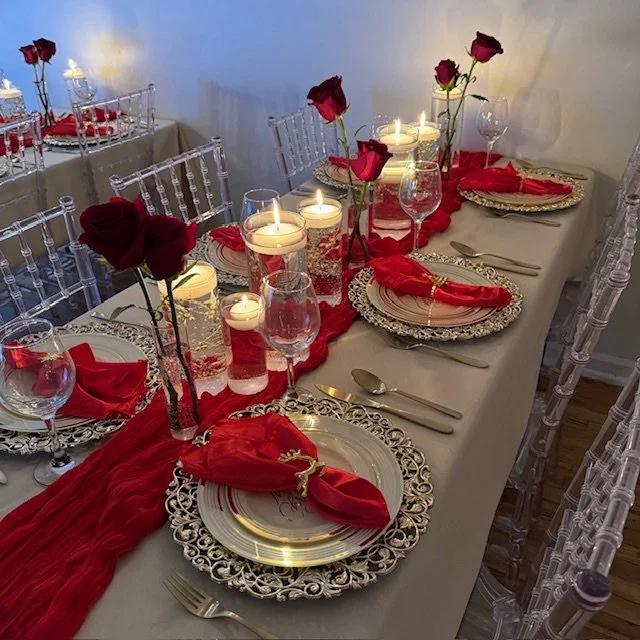 Elegant dining table decorated with red roses in tall vases, candles, gold chargers, red napkins, and clear glassware for a special occasion.