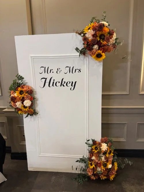 Wedding or event sign with the text "Mr. & Mrs. Hickey" decorated with arrangements of sunflowers, roses, and other flowers in orange, yellow, white, and burgundy, positioned in each corner of the white standing sign.
