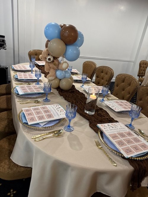 Elegant dining table decorated with a balloon centerpiece featuring blue, brown, and beige balloons, a teddy bear, and a brown table runner. Set with glassware, menus, gold flatware, and small candles for a celebration or party.