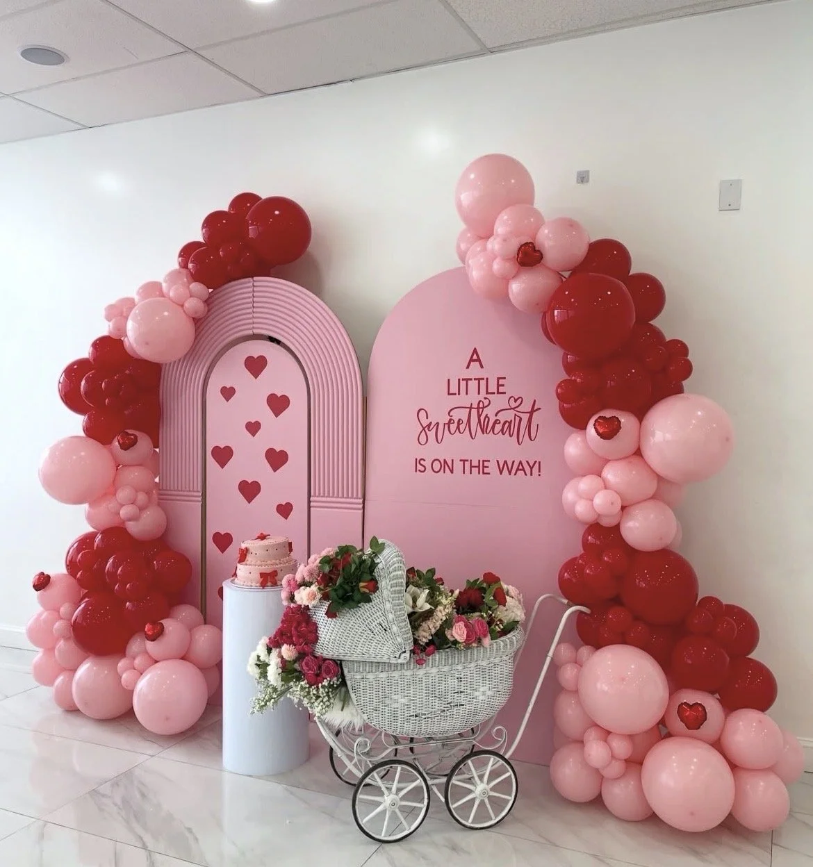 Decorative display with pink and red balloons surrounding a pink backdrop with the message 'A little sweetheart is on the way!' A white wicker baby carriage filled with pink and red flowers, a small cake, and a pink decorative arch with hearts.