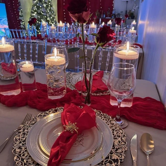 Decorated table for a romantic meal with candles, red roses, ornate plate, red napkin with gold snowflake pin, water glass, and surrounding candles with Christmas trees and additional tables in the background.