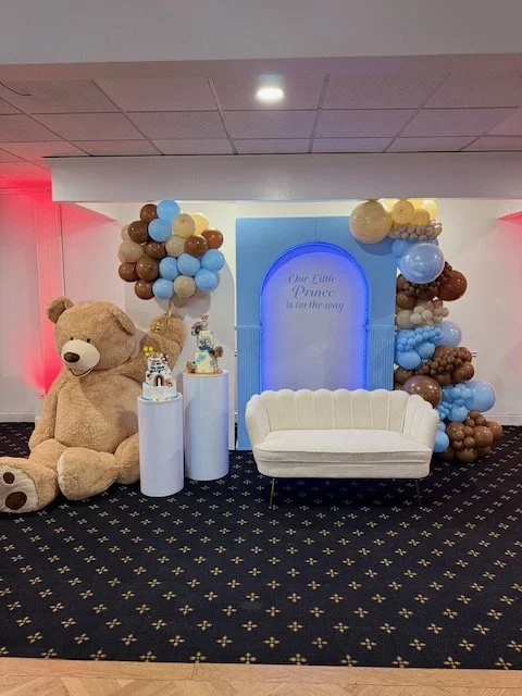 Kids' birthday party setup with a giant teddy bear, balloons, a small white sofa, and decorated tables with birthday cakes.