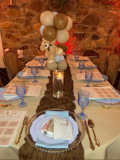 Decorated banquet table with a teddy bear centerpiece, balloons, blue glassware, menus, and place settings in a rustic setting.
