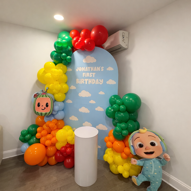 Colorful balloon decorations in red, yellow, green, orange, and blue surround a pastel blue backdrop with clouds and birthday message for Jonathan's first birthday, including decorative character balloons resembling a smiling TV and a cartoon baby in pajamas.