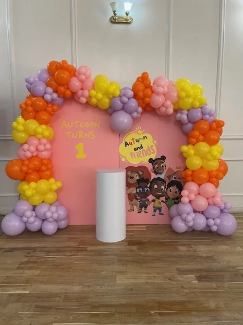 Party backdrop with colorful balloon arch in purple, pink, orange, and yellow, a pink background with the text 'Autumn Turns 1', and animated characters from Blinq and Friends. A white cylindrical stand is in front of the backdrop.