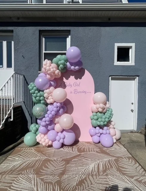 Balloons in pastel colors including pink, purple, and green arranged around a pink backdrop with text for a baby girl gender reveal event.