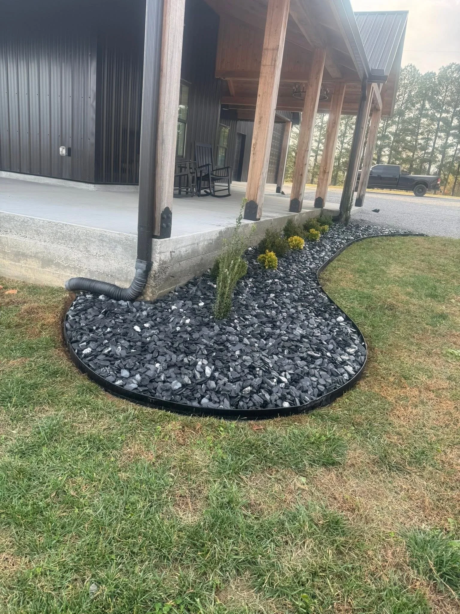 Front porch of a house with black metal siding, wooden support beams, and a concrete foundation. A small landscaped area with black mulch, small yellow bushes, and a black edging holds some plants. Green grass surrounds the landscaped patch. A gravel driveway is visible in the background, with a black pickup truck parked near the house.
