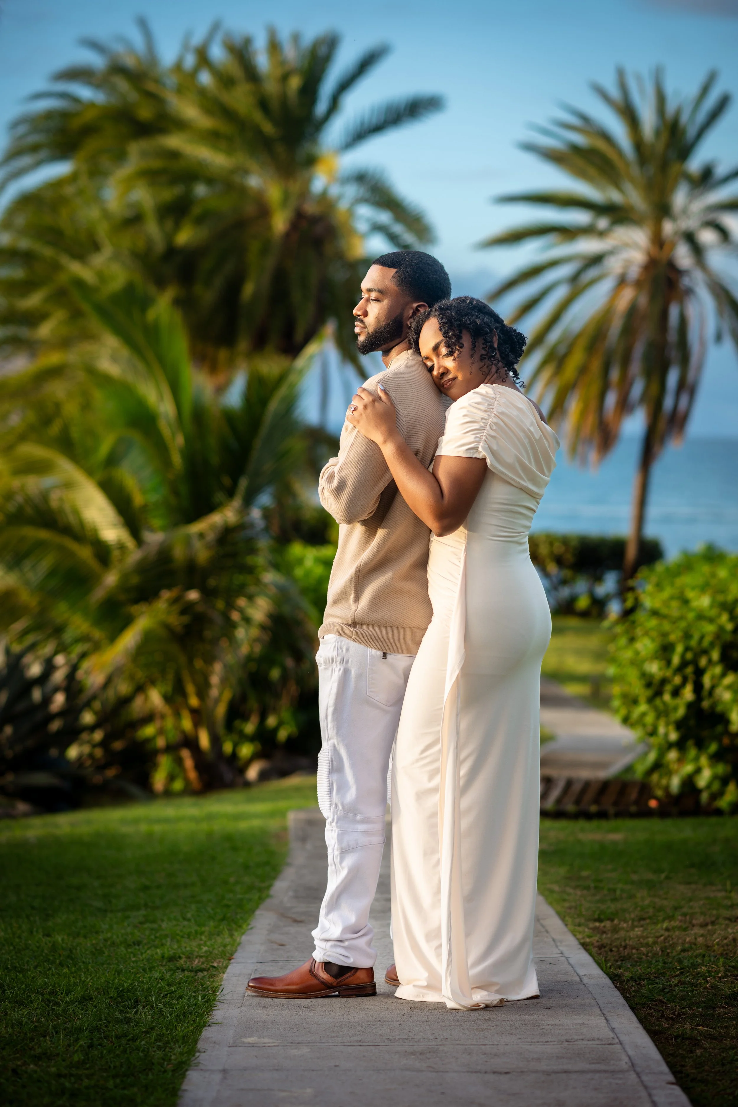 A couple hugging outdoors near palm trees, with ocean in the background, during golden hour.
