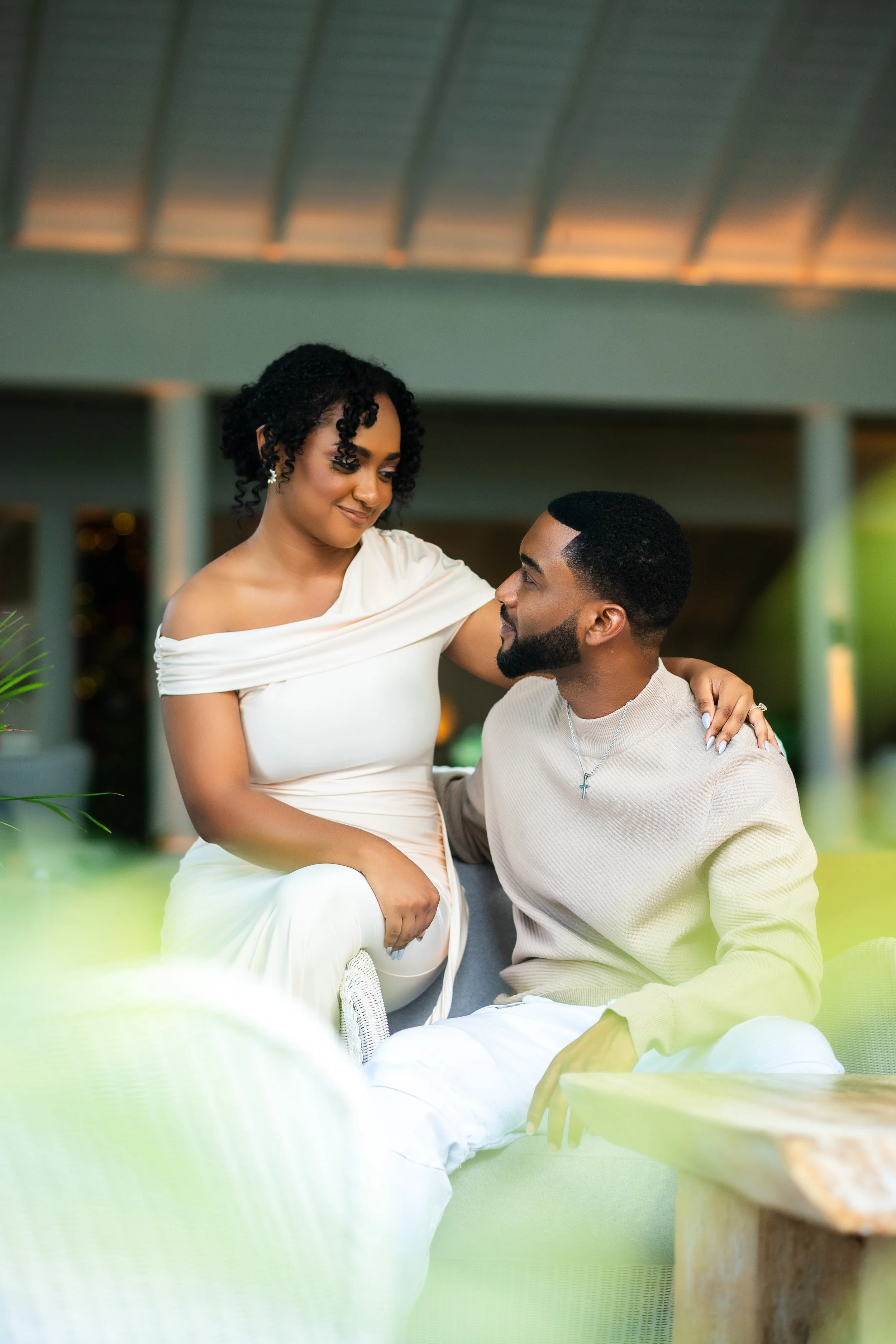 A woman in a white off-shoulder dress gently touching a man's shoulder while looking at him, both smiling warmly, sitting close together in a cozy indoor setting.