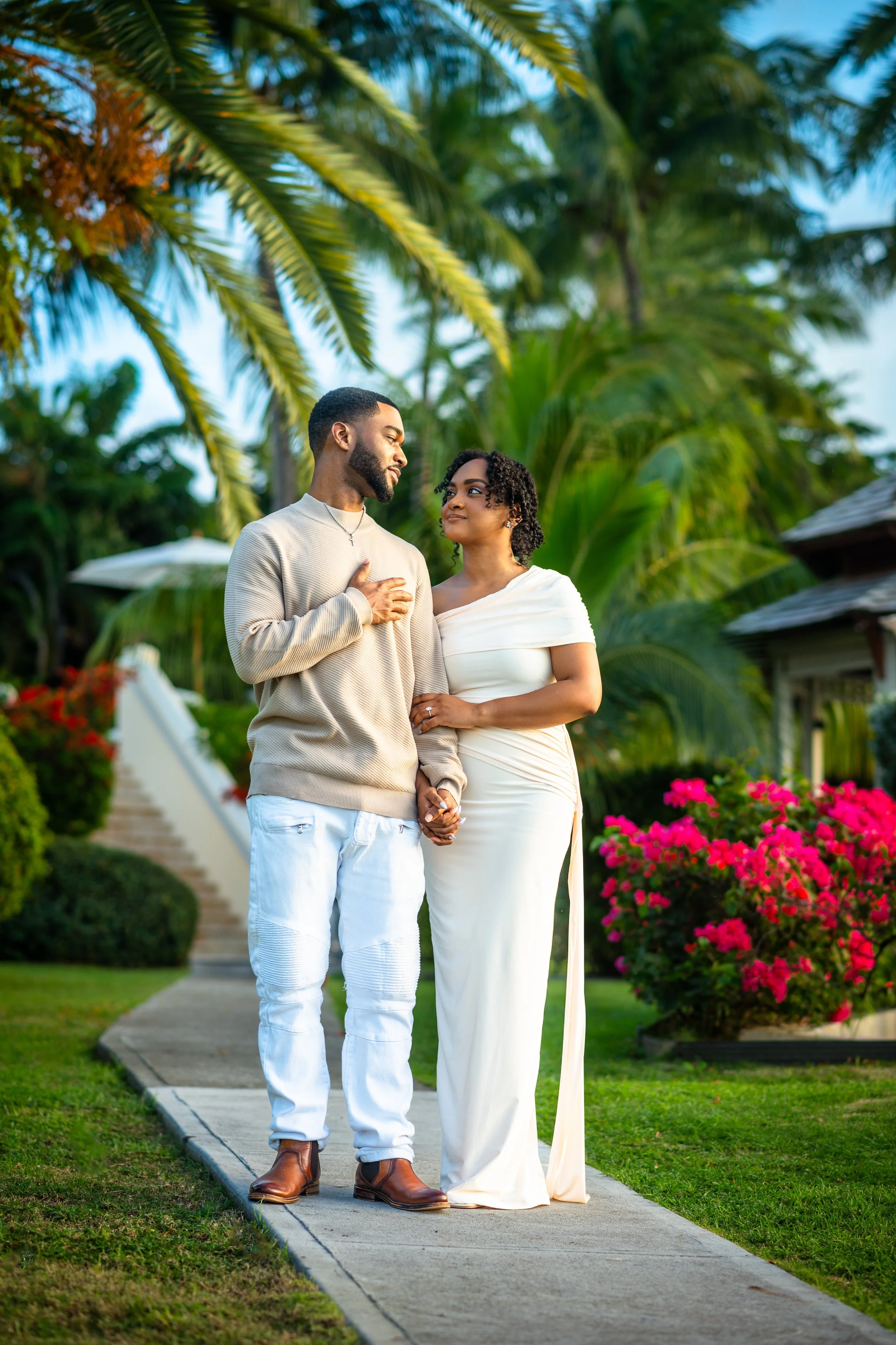 A couple standing on a garden pathway with tropical plants and pink flowers, holding hands and looking at each other romantically.