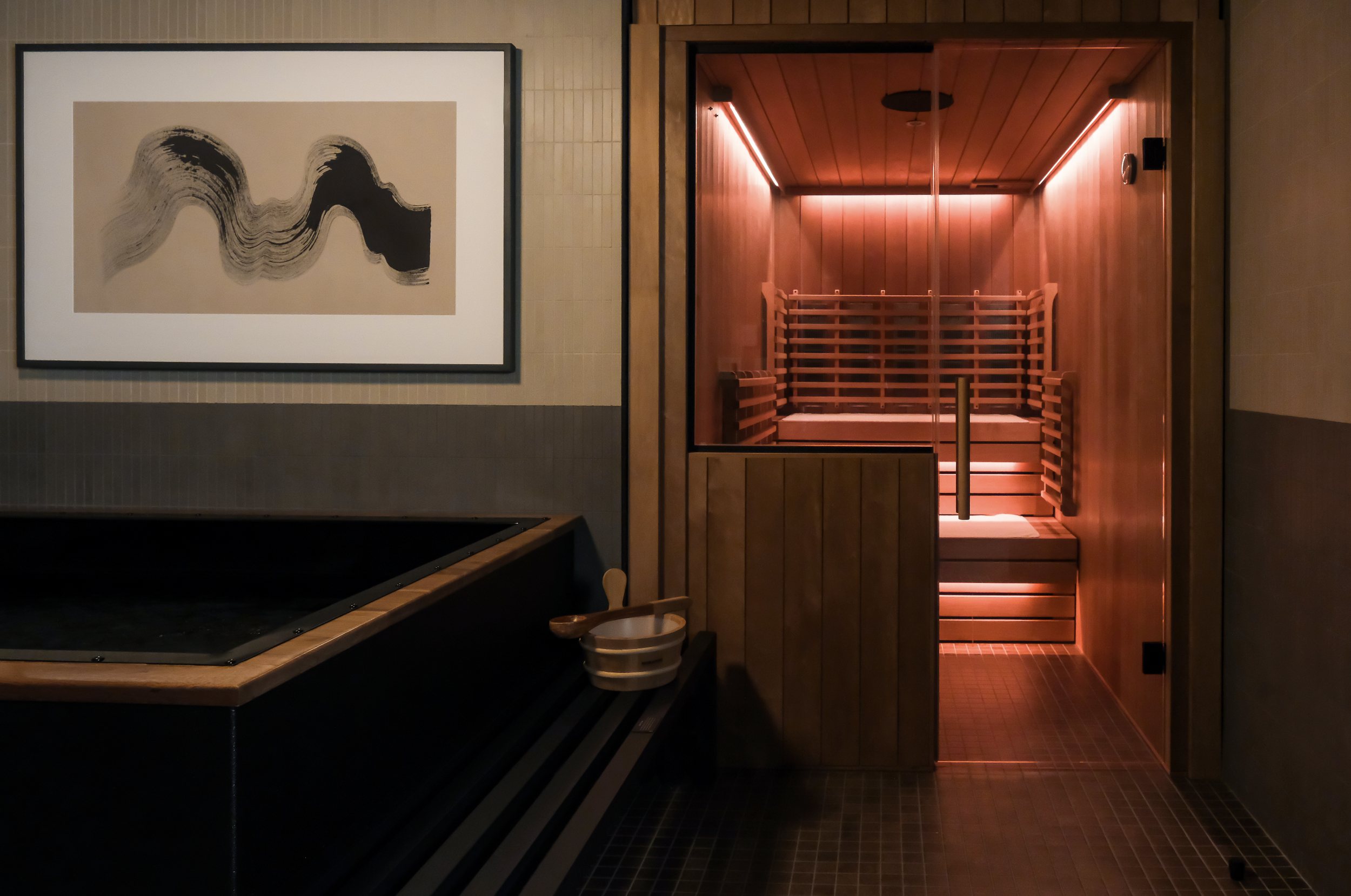 A sauna room with wooden interior and a glass door, illuminated by pink lighting, next to a tiled area with a black hot tub and a small wooden bucket and ladle.