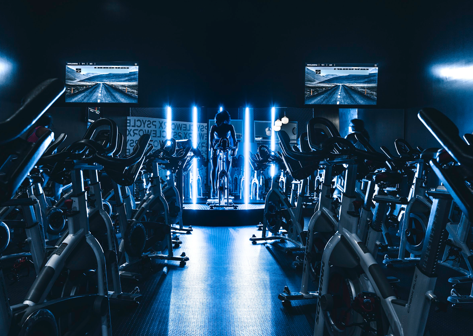 A dark indoor cycling studio with rows of exercise bikes facing large screens showing a scenic mountain road. Blue lighting highlights the equipment and a person is on a stationary bike in front of the screens.