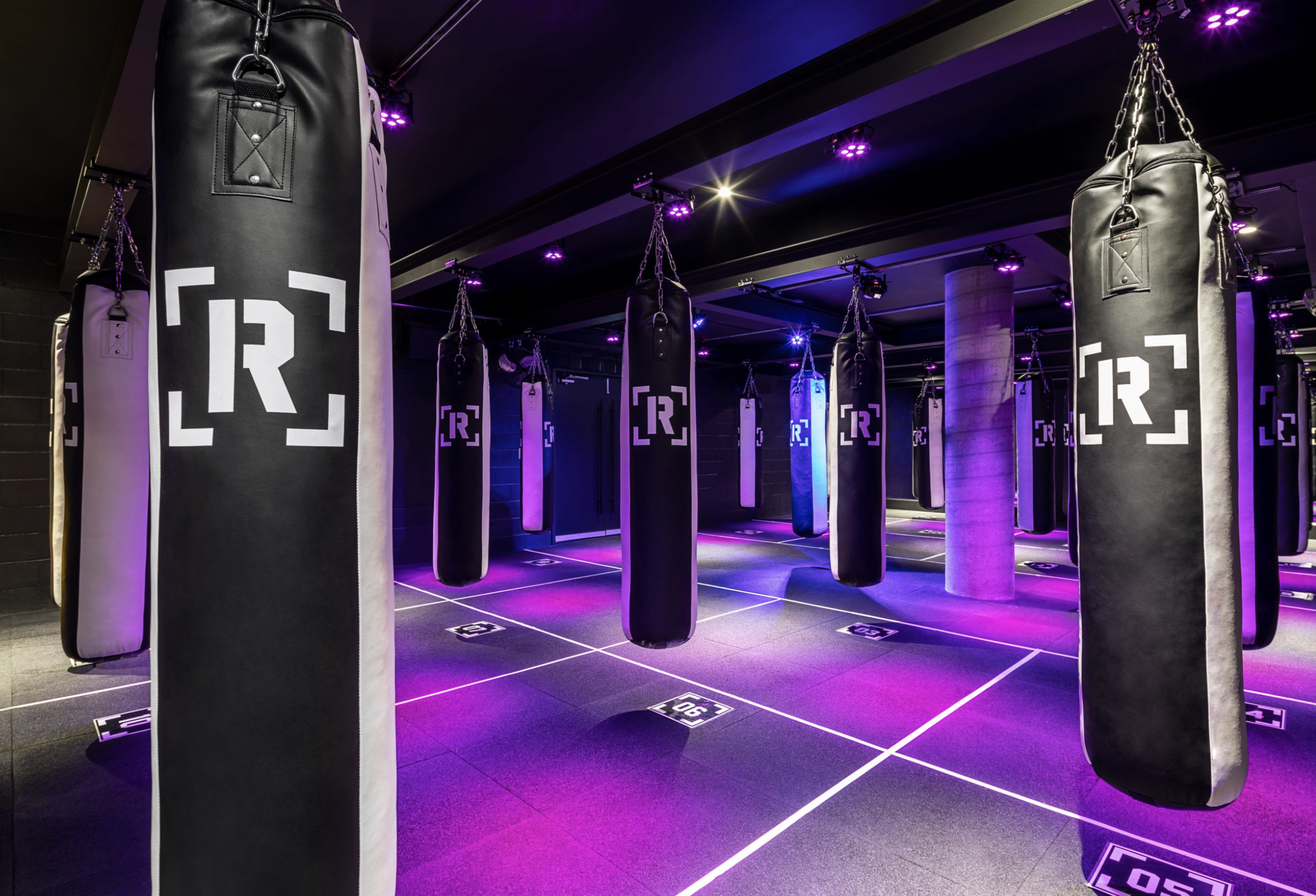 Indoor boxing gym with hanging black punching bags marked with a white logo, illuminated by purple and pink lighting.