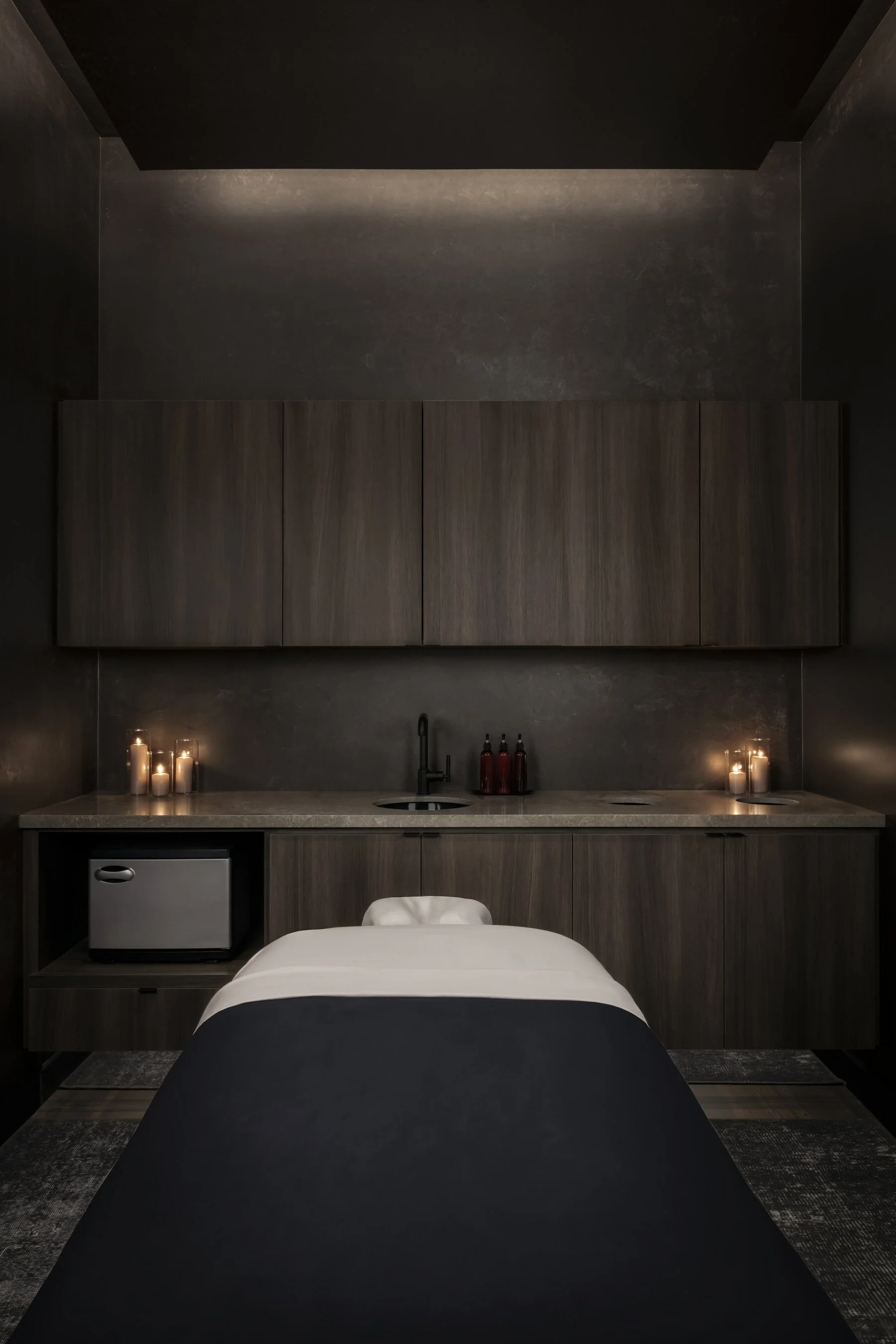 Dark, modern room with a massage bed, candles, and a kitchenette area with a small refrigerator and bottles on the counter.