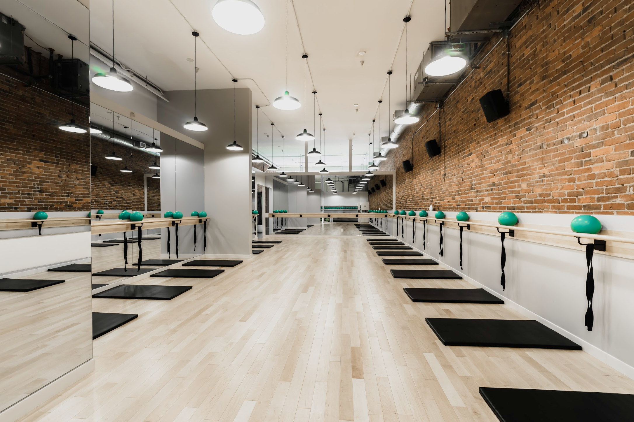 Empty fitness studio with wooden floors, brick walls, black mats, green exercise balls, and mirrors.